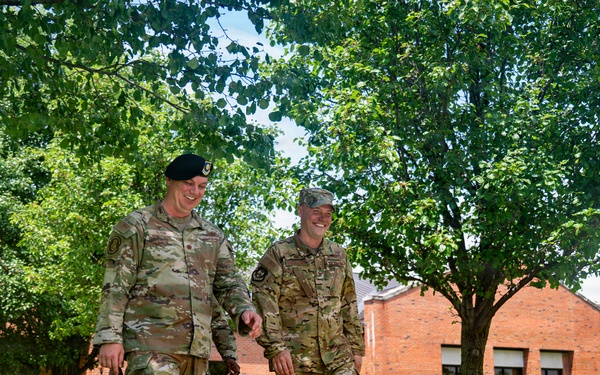Two military personnel in camouflage uniforms walk together along a paved pathway on what appears to be a military base, both smiling and engaged in conversation. The setting includes well-maintained grass, mature trees, and red brick buildings typical of a military installation. Additional uniformed personnel are visible in the background.