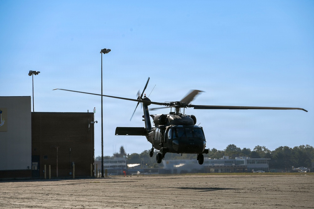 A military helicopter, consistent with a UH-60 Black Hawk, is captured in low hover or landing/takeoff phase over a paved apron at what appears to be a military airfield or joint-use airport. The rotor blades show significant motion blur, indicating active flight. The setting includes hangar buildings, light poles, and tree lines in the background under clear blue skies.