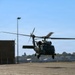 A military helicopter, consistent with a UH-60 Black Hawk, is captured in low hover or landing/takeoff phase over a paved apron at what appears to be a military airfield or joint-use airport. The rotor blades show significant motion blur, indicating active flight. The setting includes hangar buildings, light poles, and tree lines in the background under clear blue skies.