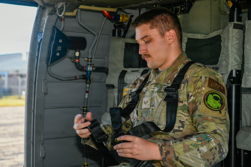 A U.S. Air Force airman in OCP (Operational Camouflage Pattern) uniform is seated inside what appears to be a military helicopter, examining or adjusting a harness/seatbelt system. He wears a unit patch on his right sleeve and rank insignia on his chest, suggesting he is an Airman First Class. The interior of the aircraft is visible with various mechanical components, tubing, and equipment panels.