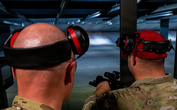 Two individuals wearing military camouflage uniforms (OCP/MultiCam pattern) are at an indoor shooting range. One person is actively aiming a firearm downrange while the other observes, consistent with a training or qualification exercise. Both are wearing red hearing protection earmuffs pushed up on their heads.