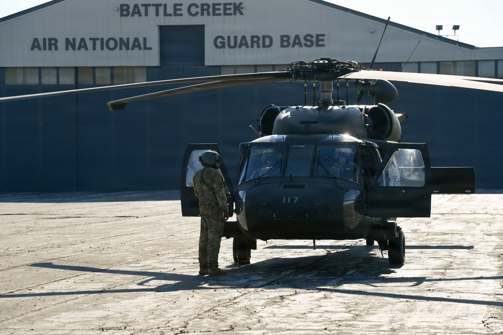 A military helicopter numbered '117', consistent with a UH-60 Black Hawk, is parked on the tarmac at Battle Creek Air National Guard Base. A soldier in camouflage uniform and flight helmet stands beside the open side door of the aircraft. The scene appears to depict pre-flight or post-flight ground operations.