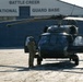 A military helicopter numbered '117', consistent with a UH-60 Black Hawk, is parked on the tarmac at Battle Creek Air National Guard Base. A soldier in camouflage uniform and flight helmet stands beside the open side door of the aircraft. The scene appears to depict pre-flight or post-flight ground operations.