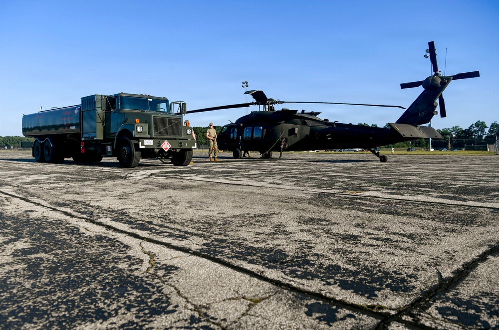 A military refueling operation is taking place on a tarmac or airfield apron. A large olive-drab military fuel tanker truck is positioned next to a UH-60 Black Hawk helicopter, with at least two personnel in military camouflage uniforms standing between the vehicles. The scene appears to be a Forward Area Refueling Point (FARP) or routine ground refueling operation.