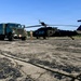 A military refueling operation is taking place on a tarmac or airfield apron. A large olive-drab military fuel tanker truck is positioned next to a UH-60 Black Hawk helicopter, with at least two personnel in military camouflage uniforms standing between the vehicles. The scene appears to be a Forward Area Refueling Point (FARP) or routine ground refueling operation.