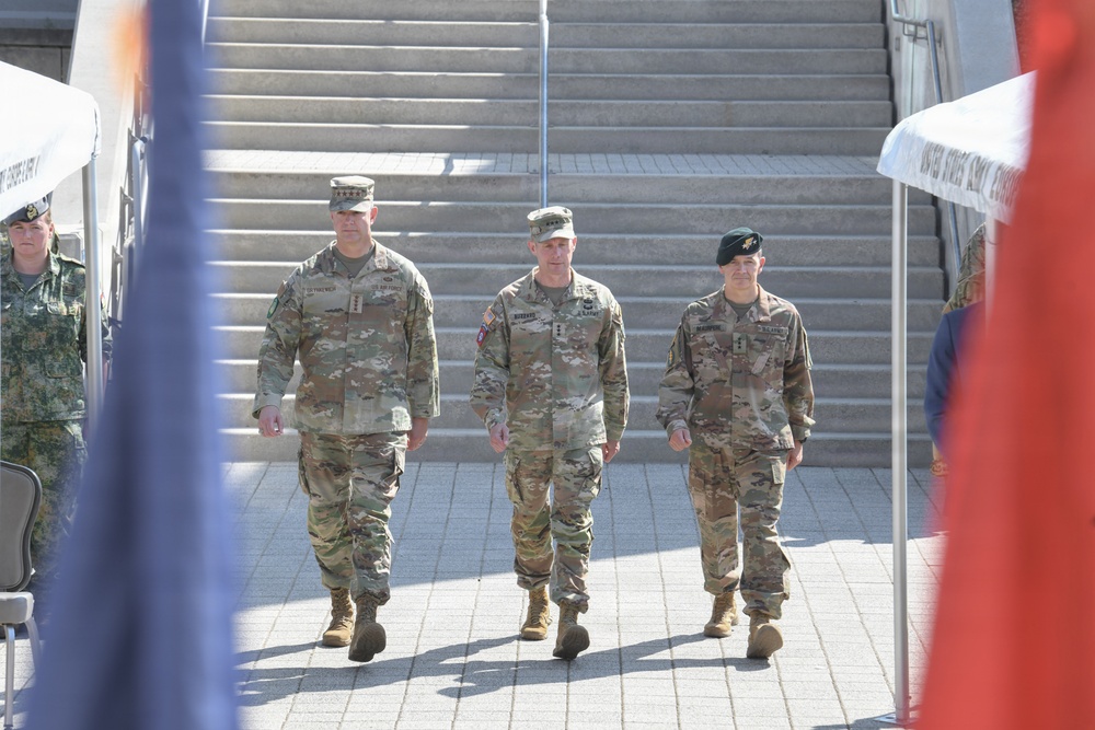 Three U.S. Army soldiers in OCP (Operational Camouflage Pattern) uniforms are walking in formation toward the camera during what appears to be a formal military ceremony or event. The soldier on the left displays four-star rank insignia on his cap, indicating a General officer. The setting includes ceremonial tents, flags, and stone steps in the background.