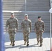 Three U.S. Army soldiers in OCP (Operational Camouflage Pattern) uniforms are walking in formation toward the camera during what appears to be a formal military ceremony or event. The soldier on the left displays four-star rank insignia on his cap, indicating a General officer. The setting includes ceremonial tents, flags, and stone steps in the background.