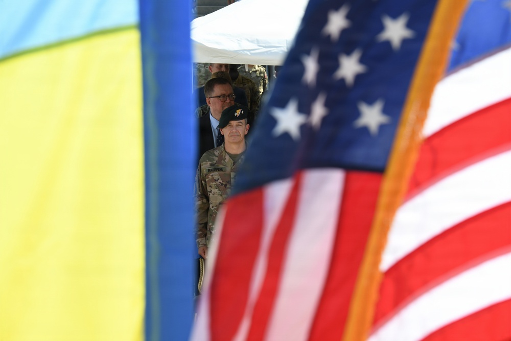 The image shows military personnel framed between a Ukrainian flag on the left and a United States flag on the right, suggesting a joint military event or ceremony. A soldier wearing a black beret and camouflage uniform is prominently visible in the center, with additional uniformed and civilian personnel visible in the background. The composition emphasizes the bilateral nature of the gathering through the deliberate framing of the two national flags.