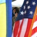 The image shows military personnel framed between a Ukrainian flag on the left and a United States flag on the right, suggesting a joint military event or ceremony. A soldier wearing a black beret and camouflage uniform is prominently visible in the center, with additional uniformed and civilian personnel visible in the background. The composition emphasizes the bilateral nature of the gathering through the deliberate framing of the two national flags.