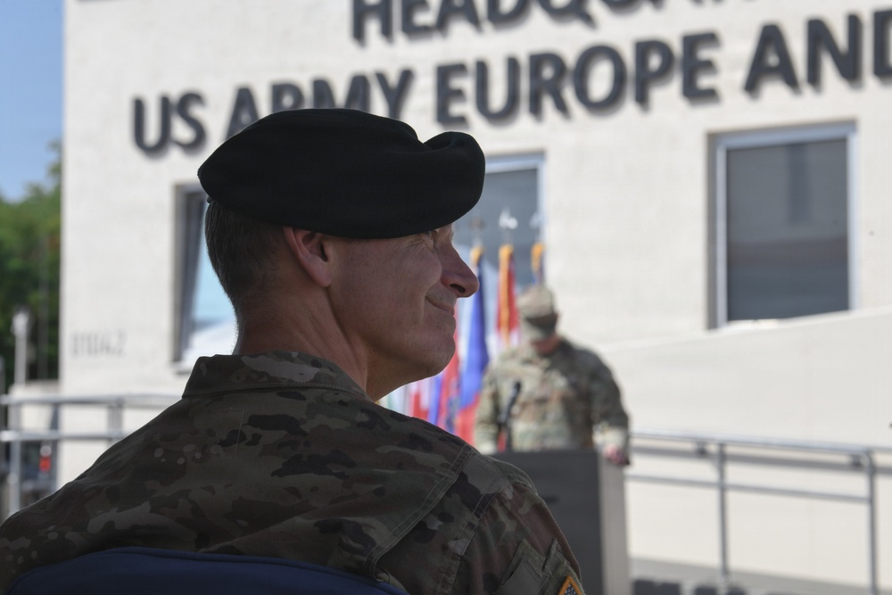 A US Army soldier wearing a black beret and multicam/OCP camouflage uniform is photographed in profile at what appears to be an official military ceremony. The setting is in front of the Headquarters, US Army Europe and Africa building. A second soldier and military color guard flags are visible in the background.