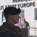 A US Army soldier wearing a black beret and multicam/OCP camouflage uniform is photographed in profile at what appears to be an official military ceremony. The setting is in front of the Headquarters, US Army Europe and Africa building. A second soldier and military color guard flags are visible in the background.