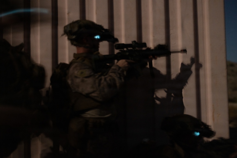 A nighttime tactical operation showing at least one soldier in full combat gear aiming a rifle through or along a metal barrier. The soldier is equipped with night vision devices mounted on their helmet, visible by the faint blue-green glow. The image is dark and partially obscured, consistent with low-light operational photography.