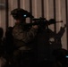 A nighttime tactical operation showing at least one soldier in full combat gear aiming a rifle through or along a metal barrier. The soldier is equipped with night vision devices mounted on their helmet, visible by the faint blue-green glow. The image is dark and partially obscured, consistent with low-light operational photography.