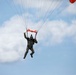 A parachutist in military camouflage uniform and helmet is descending under a partially visible red/orange parachute canopy against a partly cloudy blue sky. The jumper is holding orange marker flags or streamers in both hands, which is consistent with military or demonstration jump protocols. The image captures the skydiver mid-descent at a relatively low altitude.