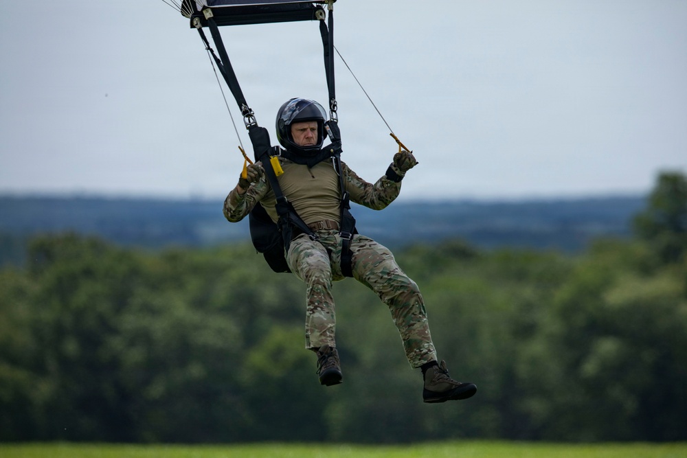A military parachutist is captured mid-descent, approaching a landing zone over a green field. The individual is wearing full military camouflage (MultiCam/OCP pattern) and a black helmet, gripping yellow toggle handles to steer the parachute canopy visible at the top of the frame. The background shows a lush treeline under an overcast sky.