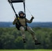 A military parachutist is captured mid-descent, approaching a landing zone over a green field. The individual is wearing full military camouflage (MultiCam/OCP pattern) and a black helmet, gripping yellow toggle handles to steer the parachute canopy visible at the top of the frame. The background shows a lush treeline under an overcast sky.