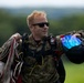 A male skydiver in camouflage military-style uniform is walking after a landing, carrying a bundled parachute with lines and a helmet with a blue-tinted visor. He is wearing black sunglasses and has a patch on his chest, suggesting affiliation with a skydiving team or military unit. The background shows a green grassy field and tree line, consistent with a drop zone or airfield.