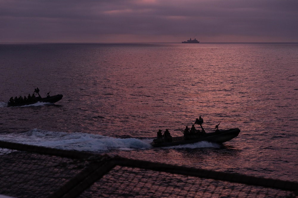 Two rigid inflatable boats (RIBs) carrying multiple personnel are underway on open water during twilight or dusk conditions. A larger vessel is visible on the horizon in the background. The image is captured from an elevated vantage point, likely from a ship, with netting visible in the foreground.