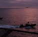 Two rigid inflatable boats (RIBs) carrying multiple personnel are underway on open water during twilight or dusk conditions. A larger vessel is visible on the horizon in the background. The image is captured from an elevated vantage point, likely from a ship, with netting visible in the foreground.