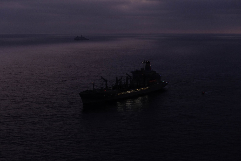 A large vessel is photographed at sea during twilight or low-light conditions, appearing as a dark silhouette against a calm ocean surface. A second, smaller vessel is visible in the distant background. The overcast sky and dim lighting create a moody, dark atmosphere across the entire scene.