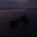 A large vessel is photographed at sea during twilight or low-light conditions, appearing as a dark silhouette against a calm ocean surface. A second, smaller vessel is visible in the distant background. The overcast sky and dim lighting create a moody, dark atmosphere across the entire scene.