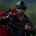 A military paratrooper, numbered '03' and wearing Polish Armed Forces insignia, is carrying a bundled red parachute after a landing. He is equipped with a tactical helmet mounted with a GoPro camera, dark sunglasses, and a full harness system. The soldier appears to be smiling or making a facial expression, suggesting a successful jump.