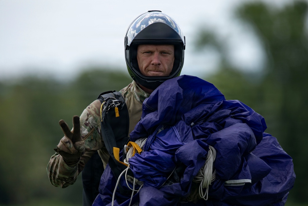 A male skydiver is shown post-landing, carrying a bundled blue/purple parachute canopy with visible lines and yellow deployment hardware. He is wearing a full-face helmet, camouflage military-style jumpsuit, and tactical gloves, and is making a peace/victory sign with his left hand. The background shows a blurred green treeline and overcast sky.