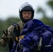 A male skydiver is shown post-landing, carrying a bundled blue/purple parachute canopy with visible lines and yellow deployment hardware. He is wearing a full-face helmet, camouflage military-style jumpsuit, and tactical gloves, and is making a peace/victory sign with his left hand. The background shows a blurred green treeline and overcast sky.