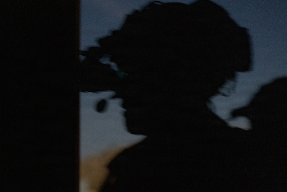 A heavily silhouetted image taken at dusk or dawn showing what appears to be a person wearing a helmet with mounted optical equipment, possibly night-vision goggles or a similar device. The figure is photographed against a dim twilight sky with blue-gray tones. A second partial silhouette is visible at the right edge of the frame.