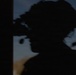 A heavily silhouetted image taken at dusk or dawn showing what appears to be a person wearing a helmet with mounted optical equipment, possibly night-vision goggles or a similar device. The figure is photographed against a dim twilight sky with blue-gray tones. A second partial silhouette is visible at the right edge of the frame.