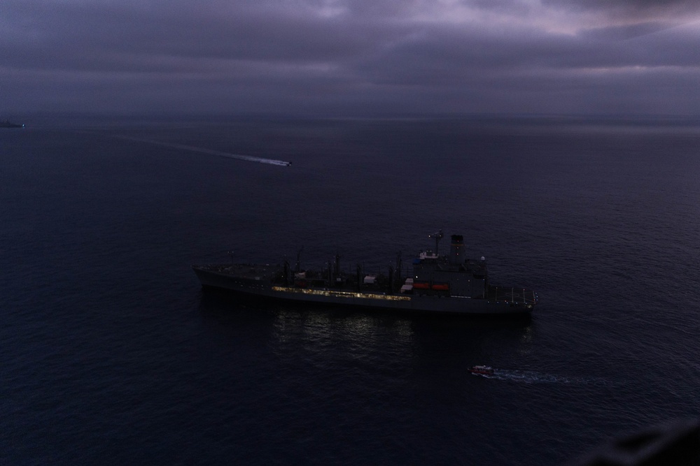 An aerial photograph taken at dusk or dawn showing a large naval or military auxiliary vessel at sea, illuminated by deck lighting against a dark, overcast sky. A smaller vessel is visible near the stern of the larger ship, and a fast-moving craft with a visible wake can be seen in the background to the left. The low-light conditions and cloud cover create a dark, moody atmosphere over calm open water.
