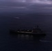 An aerial photograph taken at dusk or dawn showing a large naval or military auxiliary vessel at sea, illuminated by deck lighting against a dark, overcast sky. A smaller vessel is visible near the stern of the larger ship, and a fast-moving craft with a visible wake can be seen in the background to the left. The low-light conditions and cloud cover create a dark, moody atmosphere over calm open water.