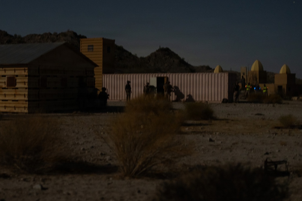 A nighttime scene showing silhouetted figures positioned around what appears to be a military training compound or mock village in a desert environment. Several structures are visible including wooden buildings and dome-topped structures resembling Middle Eastern architecture. The low-light conditions suggest either a night training exercise or operational activity.