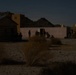 A nighttime scene showing silhouetted figures positioned around what appears to be a military training compound or mock village in a desert environment. Several structures are visible including wooden buildings and dome-topped structures resembling Middle Eastern architecture. The low-light conditions suggest either a night training exercise or operational activity.