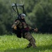 A military paratrooper is captured mid-landing on a green grassy field, still connected to a partially collapsed parachute. The individual is wearing camouflage uniform with a visible shoulder patch, a helmet with visor, and is holding yellow toggle handles used to steer the parachute. The background consists of dense green trees, suggesting a rural or training area.