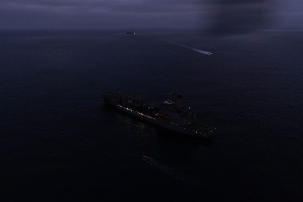An aerial nighttime photograph showing a large naval or military vessel illuminated with running lights on a dark open ocean. A second vessel or fast-moving craft is visible in the background leaving a white wake trail. The image is taken from an elevated vantage point, likely from an aircraft or drone, under overcast skies.