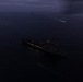 An aerial nighttime photograph showing a large naval or military vessel illuminated with running lights on a dark open ocean. A second vessel or fast-moving craft is visible in the background leaving a white wake trail. The image is taken from an elevated vantage point, likely from an aircraft or drone, under overcast skies.
