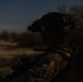 A military or law enforcement operator is photographed at dusk or nighttime wearing full tactical gear including a helmet-mounted night vision device (NVD) with visible green/teal phosphor glow from the eyepiece. The subject is facing left in profile view, appearing to be on patrol or conducting surveillance in an open, arid terrain. The low-light conditions and silhouette composition emphasize the operational nature of the scene.