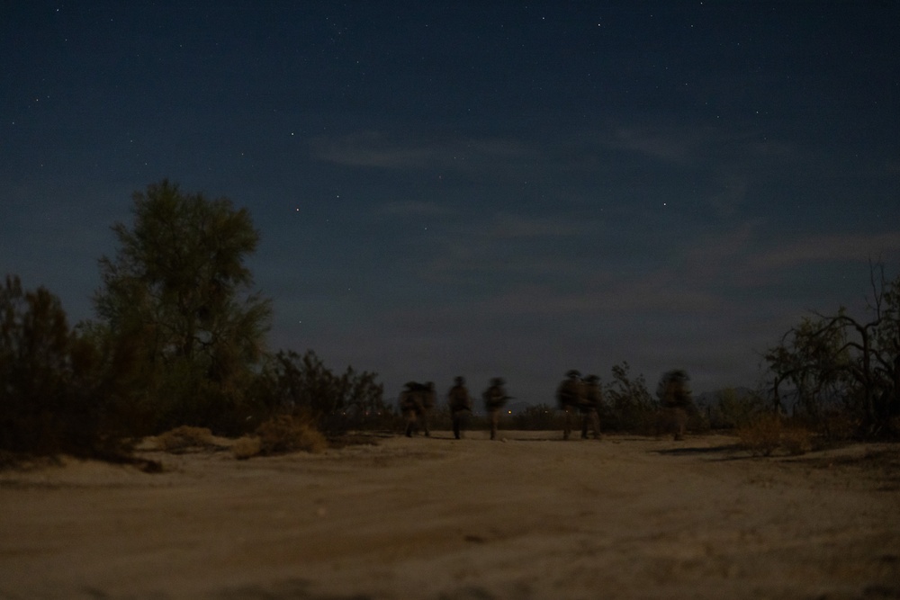 A nighttime outdoor scene showing several silhouetted figures moving across a sandy, arid landscape. The image was taken with a long exposure, causing motion blur on the figures. Stars and wispy clouds are visible in the dark blue sky above sparse desert vegetation.