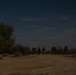 A nighttime outdoor scene showing several silhouetted figures moving across a sandy, arid landscape. The image was taken with a long exposure, causing motion blur on the figures. Stars and wispy clouds are visible in the dark blue sky above sparse desert vegetation.