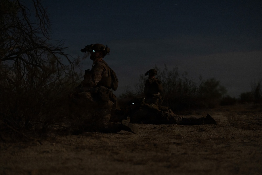A low-light nighttime image showing two military personnel in full combat gear positioned in an arid, scrub-brush terrain. The soldiers appear to be in a tactical posture, with one kneeling and one in a prone or crouched position. Night vision devices are visible mounted on their helmets.