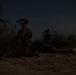 A low-light nighttime image showing two military personnel in full combat gear positioned in an arid, scrub-brush terrain. The soldiers appear to be in a tactical posture, with one kneeling and one in a prone or crouched position. Night vision devices are visible mounted on their helmets.
