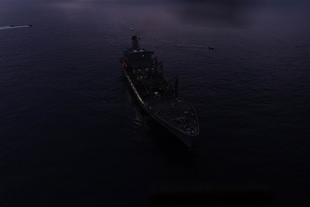 An aerial photograph taken at dusk or dawn showing a naval/military vessel at sea from a top-down perspective. The ship appears to be stationary or moving slowly, with at least one smaller vessel visible in the background. Extremely low ambient light conditions make detailed identification difficult.