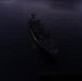 An aerial photograph taken at dusk or dawn showing a naval/military vessel at sea from a top-down perspective. The ship appears to be stationary or moving slowly, with at least one smaller vessel visible in the background. Extremely low ambient light conditions make detailed identification difficult.