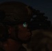 A military or law enforcement operator is photographed at night wearing a tactical helmet mounted with night vision goggles (NVG). The green-blue glow visible through the NVG eyepiece indicates the device is active and in use. The image is taken in low-light/dusk conditions, highlighting the operator's profile against a dark sky.