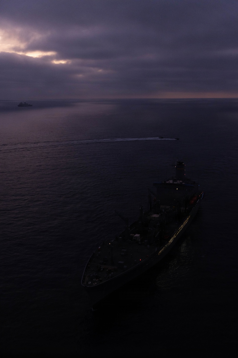 An aerial photograph taken at dusk or dawn showing a large naval or military vessel in the foreground, viewed from above and slightly ahead of the bow. A smaller vessel is visible in the middle distance leaving a white wake, and a third vessel appears on the horizon to the left. The scene is dominated by dark, overcast skies with a faint break of light near the horizon.