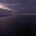 An aerial photograph taken at dusk or dawn showing a large naval or military vessel in the foreground, viewed from above and slightly ahead of the bow. A smaller vessel is visible in the middle distance leaving a white wake, and a third vessel appears on the horizon to the left. The scene is dominated by dark, overcast skies with a faint break of light near the horizon.