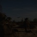 A nighttime military operation showing armed personnel on the ground with at least two helicopters visible in the sky, identifiable by their green navigation lights. The scene appears to be a tactical insertion or extraction operation in an arid, sparsely vegetated terrain. Extremely low ambient light makes fine details difficult to discern.