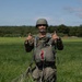 A military paratrooper stands on a grassy landing zone giving two thumbs up after a successful parachute jump. The individual is wearing full parachute gear including a helmet, harness, and reserve parachute, with the deployed canopy and suspension lines visible on the ground behind them. The setting appears to be a military drop zone during daytime with clear skies.