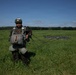 A military paratrooper stands in an open green field after a landing, holding parachute suspension lines in one hand. The soldier is wearing full combat gear including a tactical helmet with a mounted device, camouflage uniform, harness, and multiple equipment pouches. A collapsed dark parachute canopy is visible on the ground behind them.